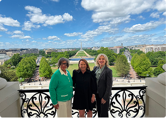 Three women standing on a balcony with a cityscape and park in the background.